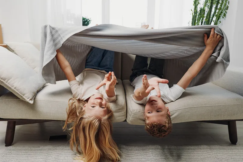 Two playful children lie upside down on a sofa covered with a blanket in a bright living room with cozy pillows and plants. Concept of childhood fun, family moments, and home comfort.