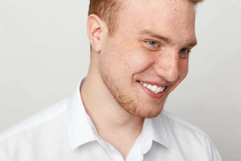 Smiling young man with red hair and freckles wearing white shirt looks down with happy expression. Close-up portrait on light background. Concept of positive emotions and natural beauty.