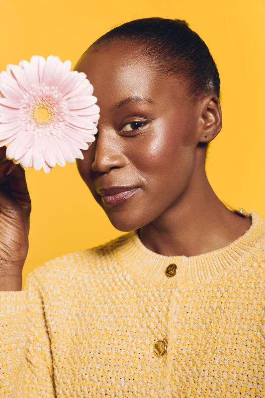 Portrait of a smiling black African woman with dark skin holding a pink flower near her face against a yellow background. Concept of beauty, natural skin, and floral wellness.