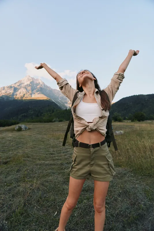 Young woman embraces outdoor freedom, raising arms in triumph against mountains, standing in an open field with a clear sky, expressing carefree lifestyle and travel vibe