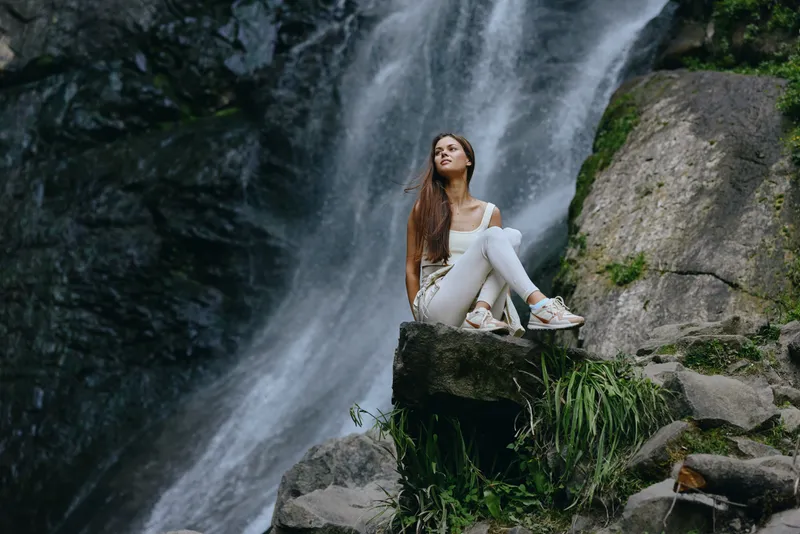 young woman sitting on rock near waterfall in nature, wearing casual white clothes, relaxed pose, outdoor adventure, scenic landscape background