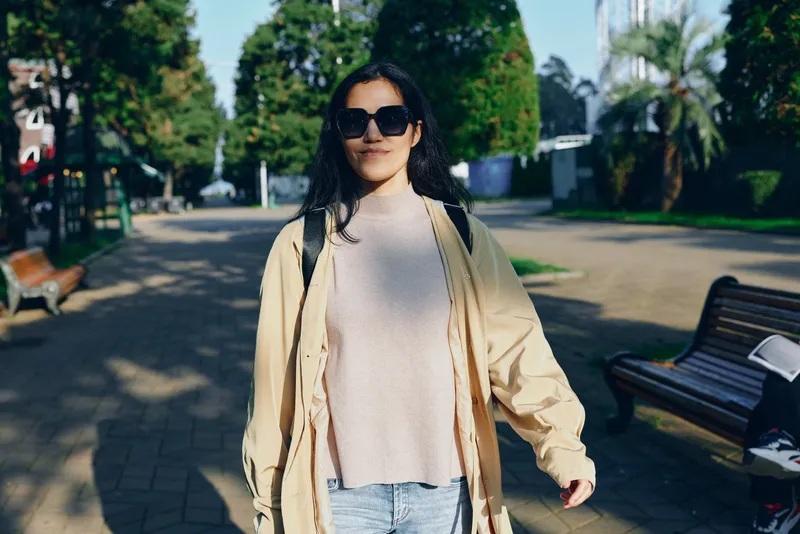 Woman outdoor fashion scene wearing a cream coat, cardigan, sunglasses, and backpack strolling along a sunny park path, conveying casual style and relaxed mood in daytime light.