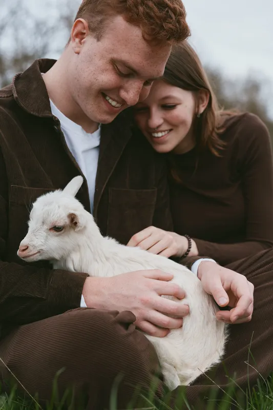 A couple in casual clothes gently petting a white goat kid outdoors, sharing a tender moment of joy in a rural setting, a scene emphasizing connection, nature, and farm life.