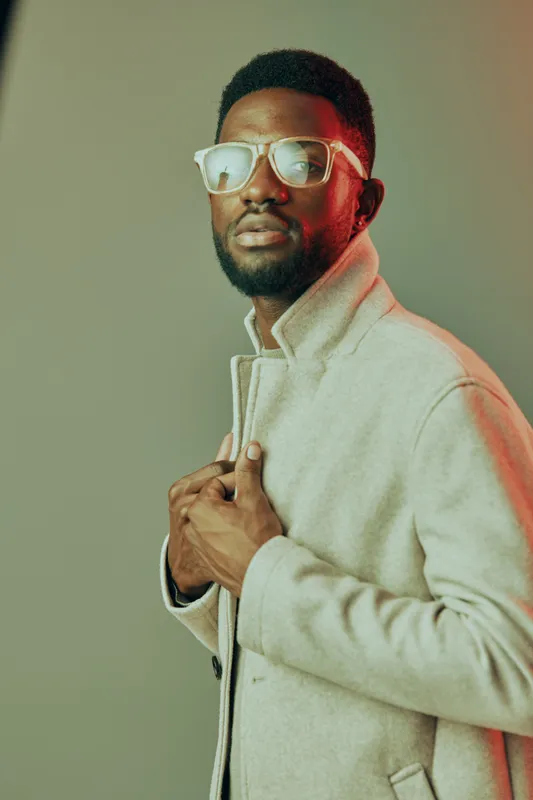 Stylish African American man wearing a trendy coat and sunglasses, posing confidently against a soft green background, showcasing modern fashion and elegance