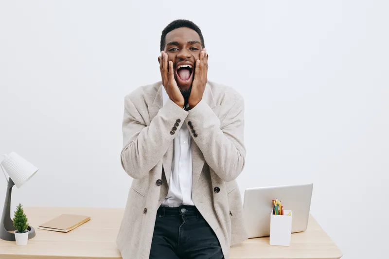 Joyful young African American man in casual clothing expressing excitement and surprise in a modern office setting with a white wall background, open laptop, and stationery decor. People lifestyle con