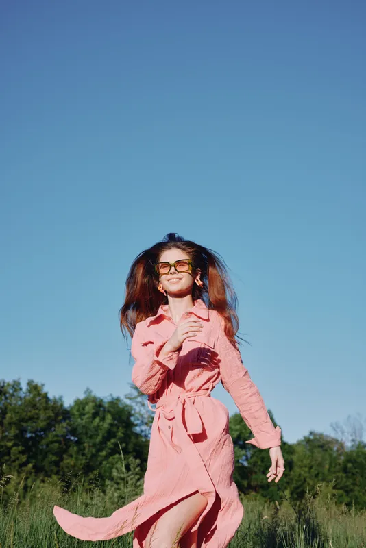 Cheerful woman in pink dress running in a sunny field, showcasing playful emotions and vibrant colors against a clear blue sky