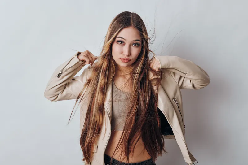 Woman with long hair and beige jacket adjusting outerwear indoors, posing with confident expression against plain white background in casual fashion style.