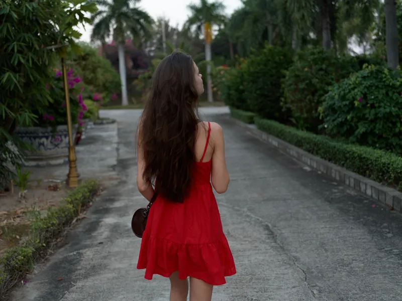 young woman with long hair wearing a red dress walking on a quiet path surrounded by greenery and palm trees, captured from behind in soft natural light