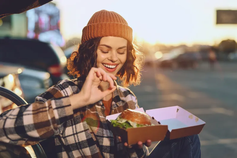 woman burger takeout smile outdoor beanie enjoying candid lifestyle moment with authenticity and golden hour glow, emotional storytelling of mindful living and urban street food joy