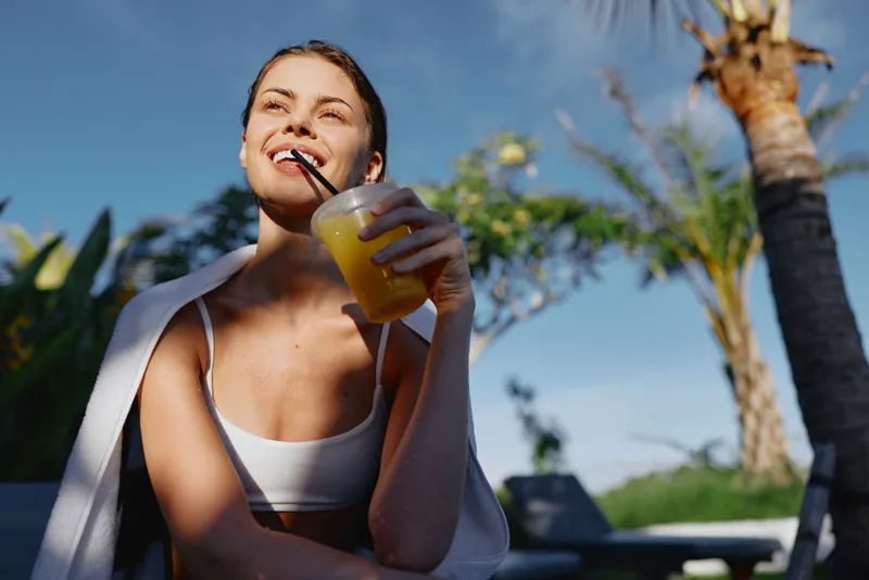 Bright happy woman enjoys juice outdoors poolside, smiling with straw in glass, wearing a white tank top, relaxed casual summer moment among palm trees and blue sky