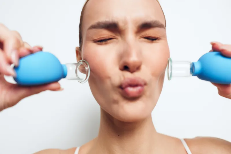Young woman using blue facial suction cups on cheeks for skin care treatment. Close-up with closed eyes on white background. Concept of beauty, wellness, and facial therapy.