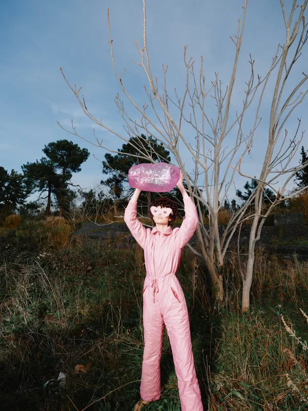 Person in pink jumpsuit holding a pink frisbee overhead in an outdoor garden with bare trees, playful fashion and sunny whimsical mood with bright color.