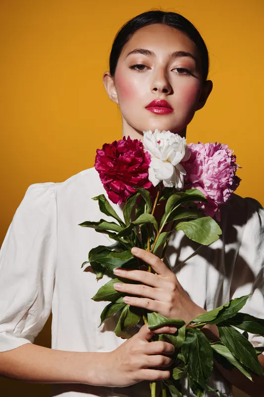 Young woman with rose makeup holds bright pink and white peonies against an orange background. Creative fashion beauty portrait with vibrant colors and soft light.