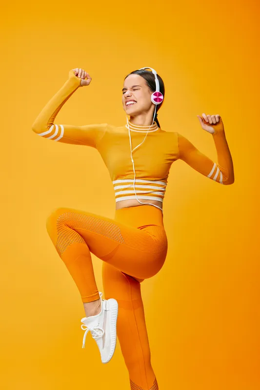 Woman in orange fitness outfit jumps with arms raised, wearing headphones, energetic workout pose against a vivid orange background, sport style, dance vibes.