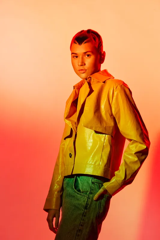 woman in yellow jacket with green pants posing confidently against warm orange background with dramatic lighting and stylish modern look