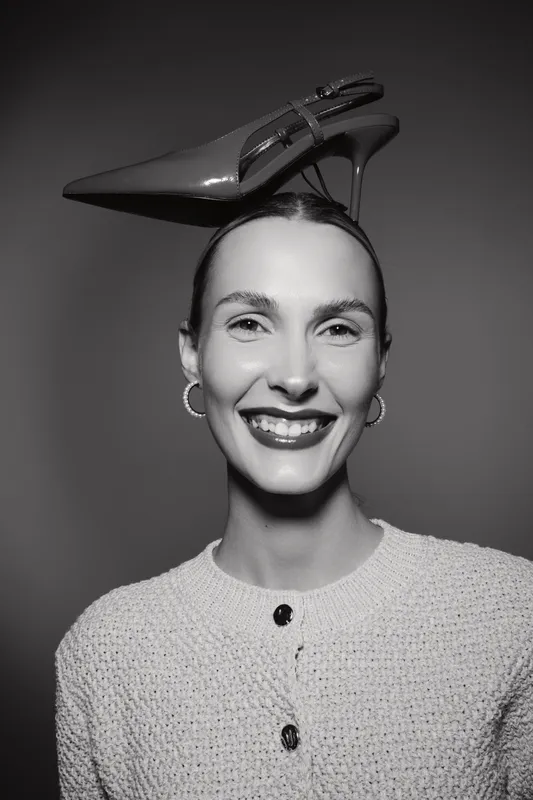 Smiling Caucasian woman with light skin and earrings balances pointed high heel shoe on head in studio. Concept of fashion, humor, and confidence.