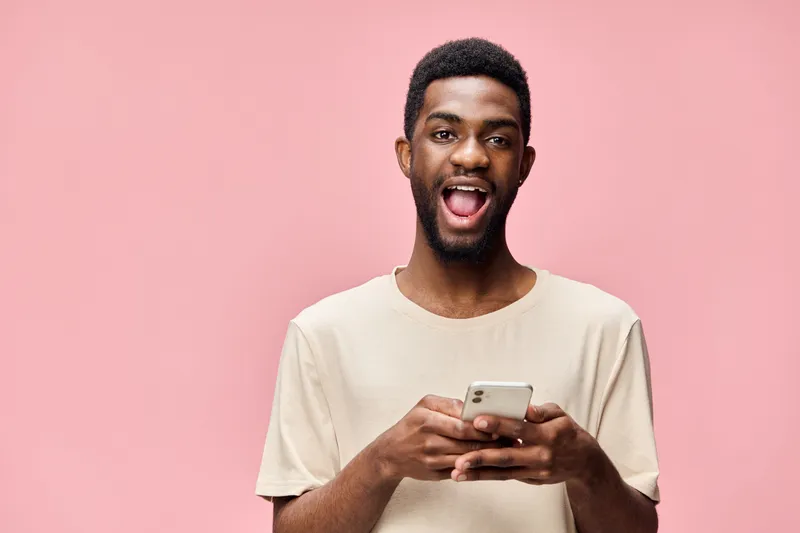 Smiling African American man using smartphone on pink background, showcasing happiness, casual style, and modern technology in a vibrant setting