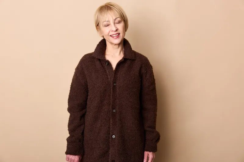 Elderly woman indoors wearing a dark brown coat, eyes closed smiling gently, relaxed posture, studio light, beige backdrop, calm mood portrait, warm soft lighting