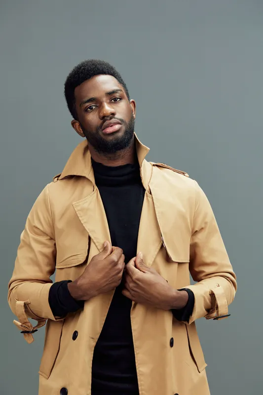 Stylish young African man in beige trench coat with black turtleneck, posing confidently against a gray background, showcasing modern fashion and self expression