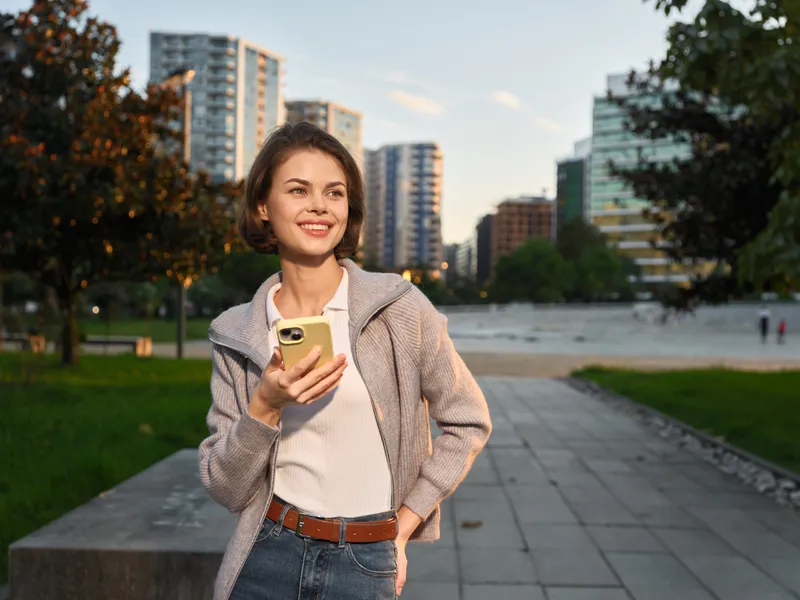 A cheerful professional woman is seen outdoors in a park, texting on a mobile device near a modern cityscape. She smiles brightly, exuding a positive and dynamic vibe.