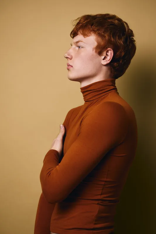 Young man with red hair wearing brown turtleneck standing sideways with arms crossed against beige background in studio portrait profile view.