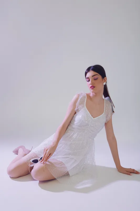 Young woman in a white lace dress poses sitting on the floor against a plain background. Concept of beauty, fashion, and elegance with soft lighting and delicate textures.
