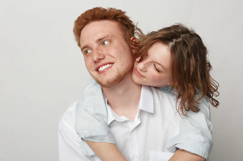 Happy young couple in casual clothes embraces and smiles on light background. The woman hugs the man from behind showing affection and warmth. Concept of love, relationship, and happiness.