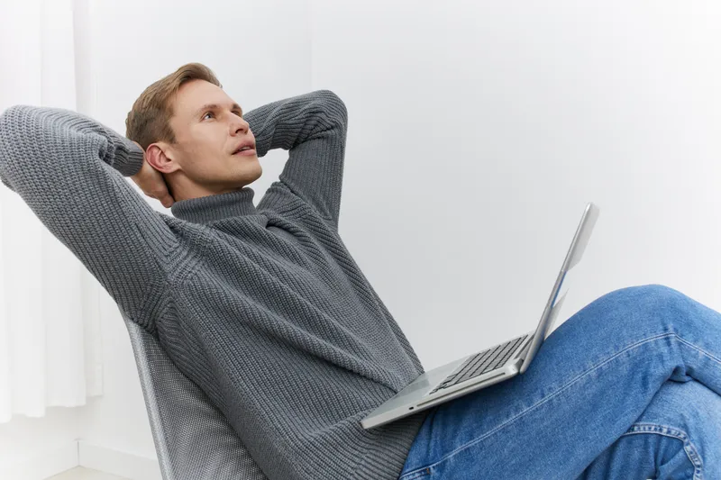 Casual young man with light brown hair wearing gray sweater and blue jeans relaxing on chair with laptop, looking upwards with thoughtful expression in bright modern home interior, lifestyle concept, 