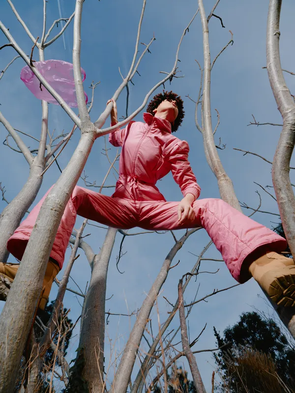 Adventure seeker in a pink jumpsuit climbs leafless trees against a blue sky, dynamic pose and airborne perspective, energetic outdoor activity.