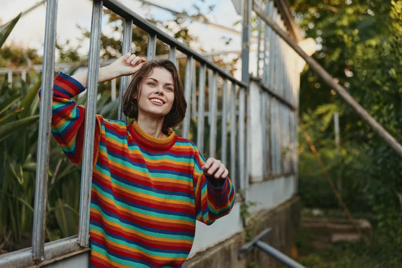 Woman portrait in greenhouse wearing colorful striped sweater, smiling and leaning on glasshouse frame in garden, casual outdoor scene with plants and natural light.