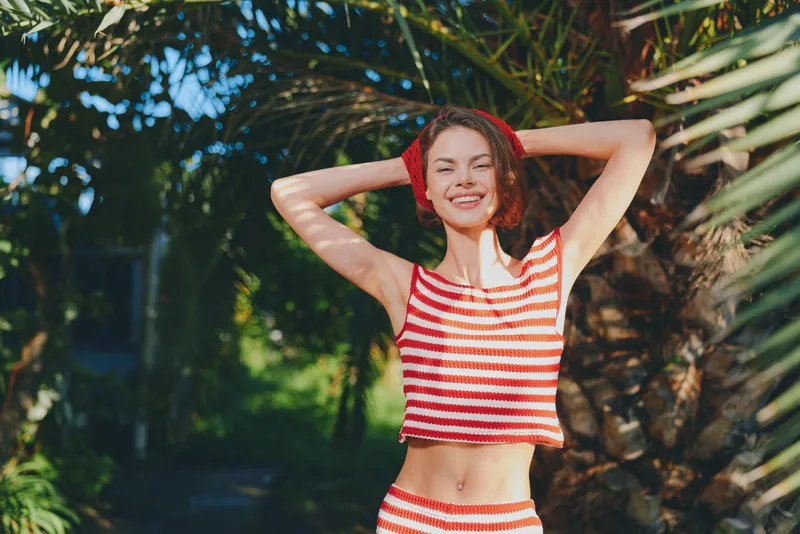 A cheerful woman wearing a striped crop top stands outdoors in a sunny garden, smiling with arms raised behind her head, conveying vitality, confidence, and a carefree, fresh summer mood.