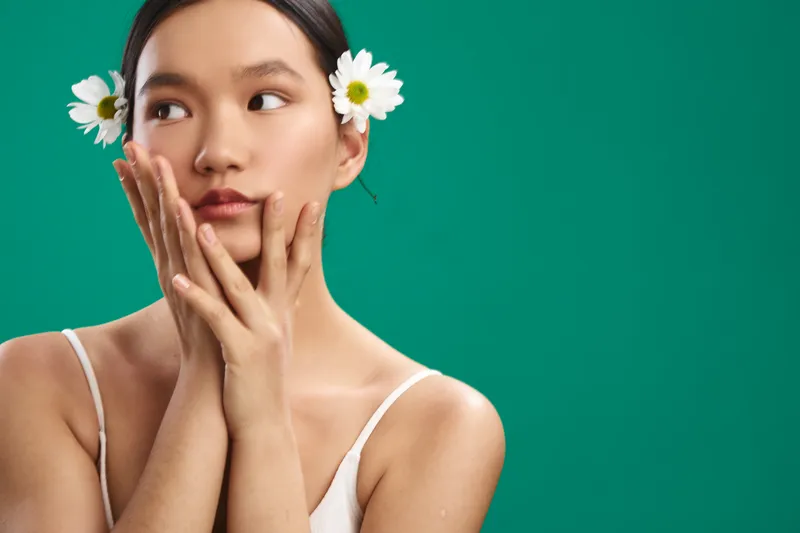 asian woman with daisies behind her ears rests her chin on her hands in a clean studio setup, conveying calm beauty and a gentle, natural vibe against a vibrant green backdrop.