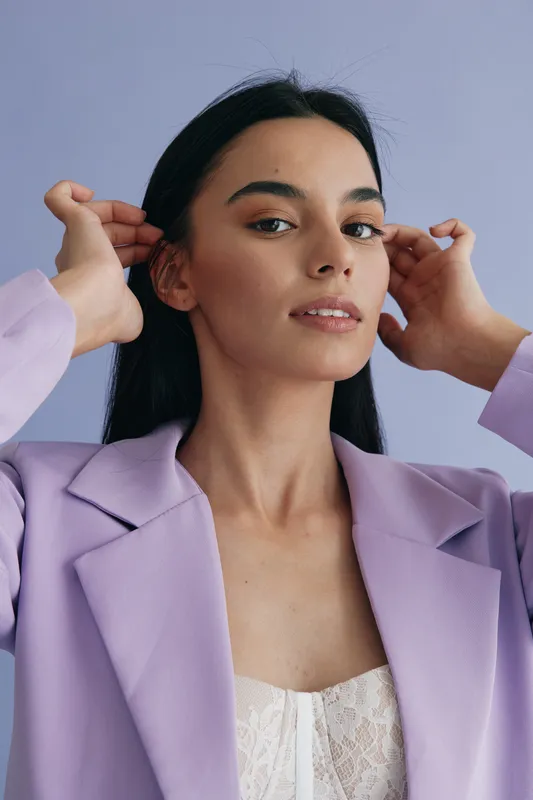 Young woman adjusting her long black hair wearing a lavender blazer and white lace top, posing confidently against a soft blue background in a minimalistic studio setting.