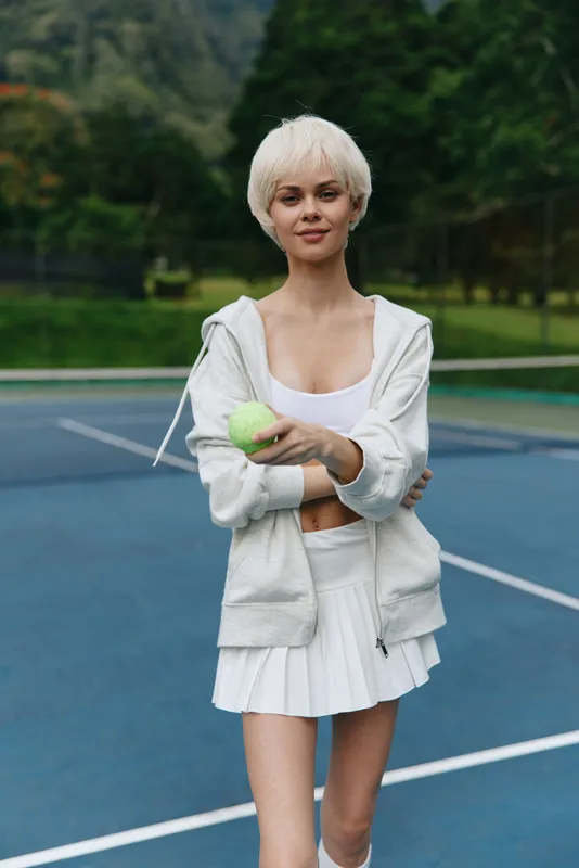 Young female tennis player in white sportswear, smiling confidently while holding a tennis ball on a vibrant blue court surrounded by greenery