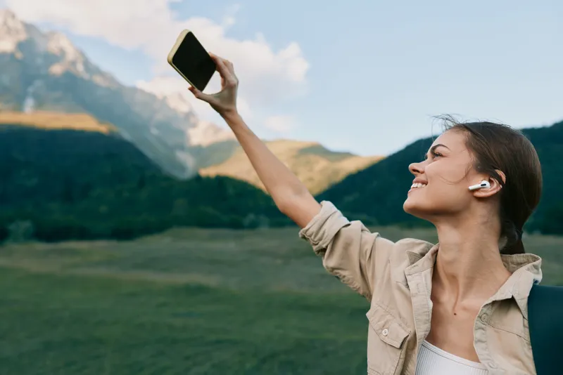 A woman takes a confident selfie in a bright mountain meadow, wearing a beige jacket and backpack, headphones on, enjoying the fresh air and expansive scenery.