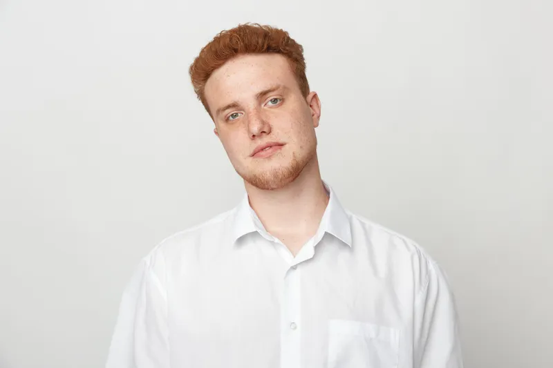 Young man with red hair and beard wearing white shirt looks at camera with neutral expression on light grey background. Concept of casual style, portrait, and natural appearance.