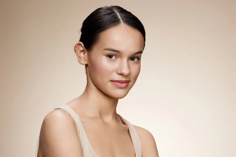 Portrait of a young woman with a natural look, wearing neutral clothing, showcasing healthy skin and a confident expression with a warm background