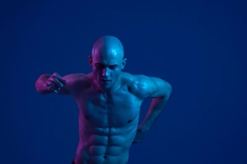 Athletic man in motion with a dynamic pose, showcasing his muscular physique against a blue background with vibrant light colors. This image captures energy and strength.