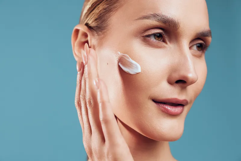 Closeup of woman applying cream on face, smooth skin texture, gentle skincare routine, natural beauty, soft lighting, blue background, focused expression, healthy complexion.