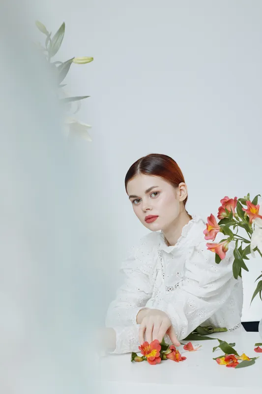 A young woman of Eastern European descent in a white blouse, sitting with colorful flowers, exuding a calm and reflective mood in a minimalist setting