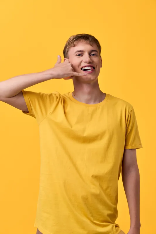Cheerful man in a yellow shirt makes a phone gesture, depicting casual communication, friendly vibe, and sunny energy against a bright studio background.