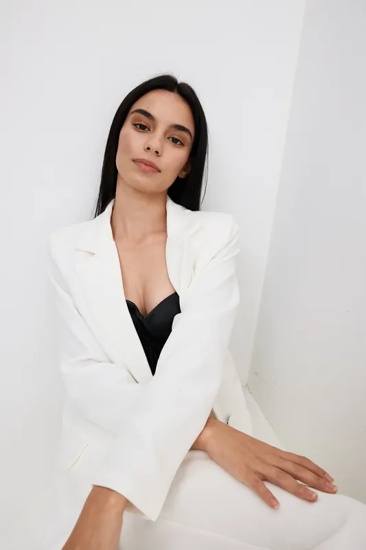 Young woman in white suit and black top posing confidently against a plain white background with a calm and composed expression, showcasing modern fashion style.