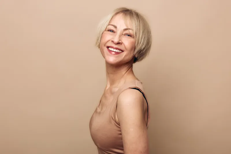 Smiling mature woman with short blonde hair wearing a sleeveless top poses confidently against beige background. Concept of happiness, aging gracefully, and natural beauty in portrait.