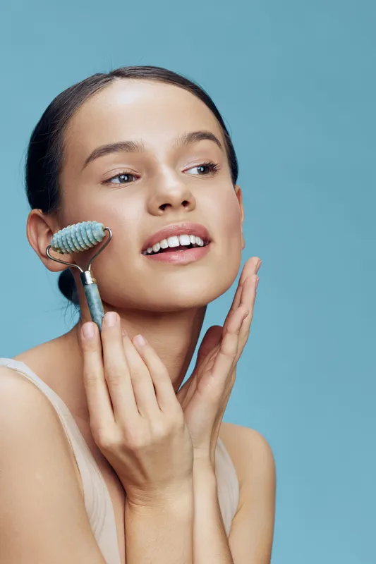 Young woman using a jade roller for skincare on a light blue background, showcasing a fresh and radiant complexion while promoting self care and beauty routines