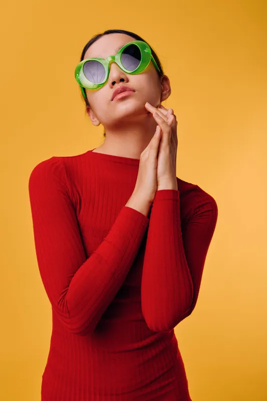 Young woman wearing green sunglasses and a red sweater poses with hands near face against orange background. Fashion, style, and confident look with modern sunglasses and casual outfit.