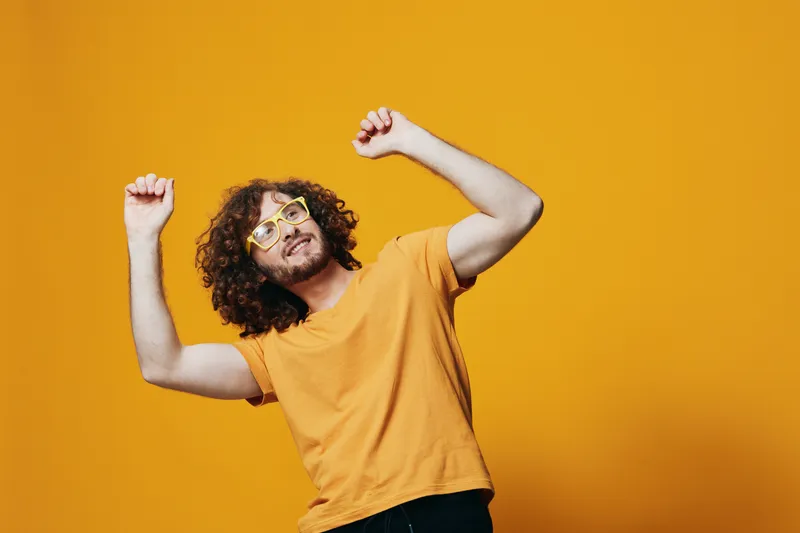 Happy young man with curly hair dancing joyfully in a bright yellow shirt and quirky glasses against a vibrant yellow background