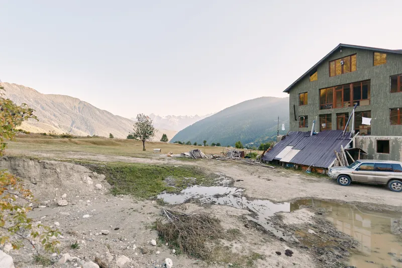 House and mountain backdrop with car parked on muddy road beside a field and small construction site in countryside, puddles and rough terrain framing a remote rural landscape.