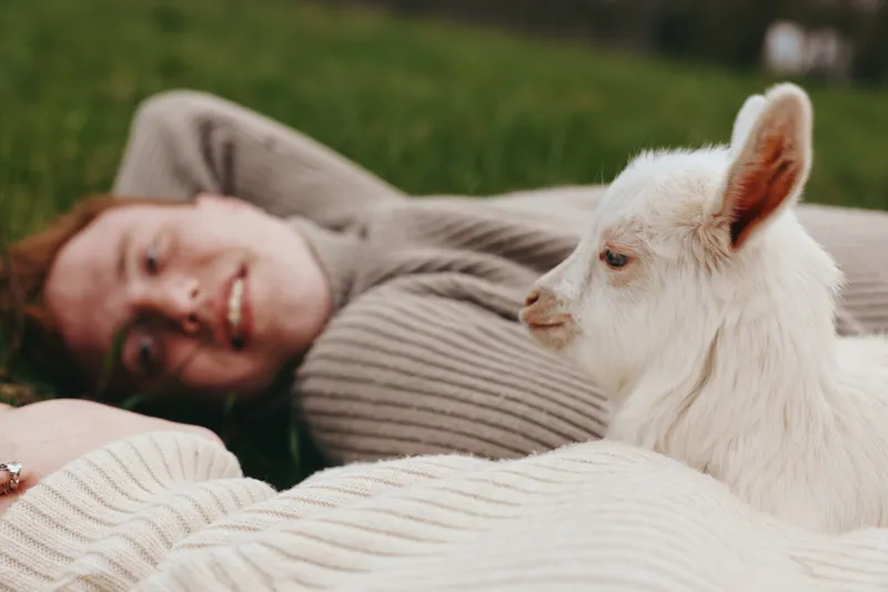 A woman relaxing on a grassy lawn with a white goat kid nearby, enjoying a peaceful moment. A candid lifestyle scene showing joy, companionship, and the simple pleasures of outdoor life.