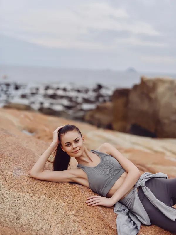 Young woman in sports attire lounging on a rocky beach, exuding confidence and calmness, perfect for wellness and fitness themes