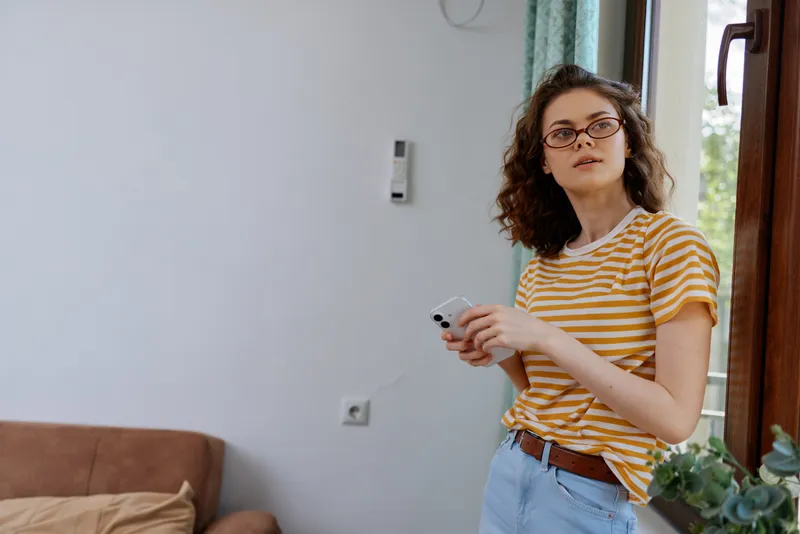 A woman in a striped t shirt stands in a bright living room at home, holding a smartphone and glancing toward the camera with a relaxed expression. Everyday home lifestyle moment.